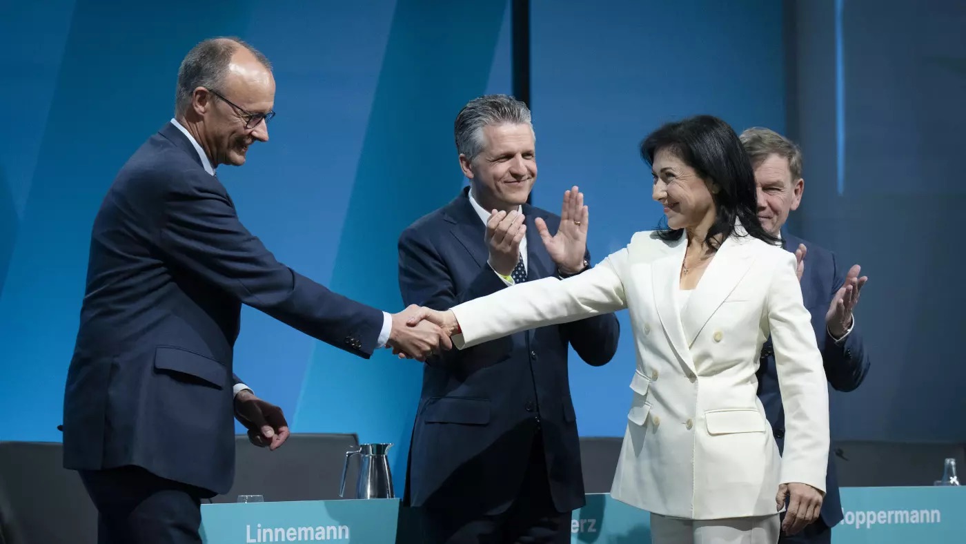 Katherina Reiche in a white suit shaking hands with Friedrich Merz on a stage in front of a blue backdrop