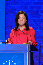 Katherina Reiche in a red jacket speaking at a lectern with a European flag motif, in front of a blue backdrop