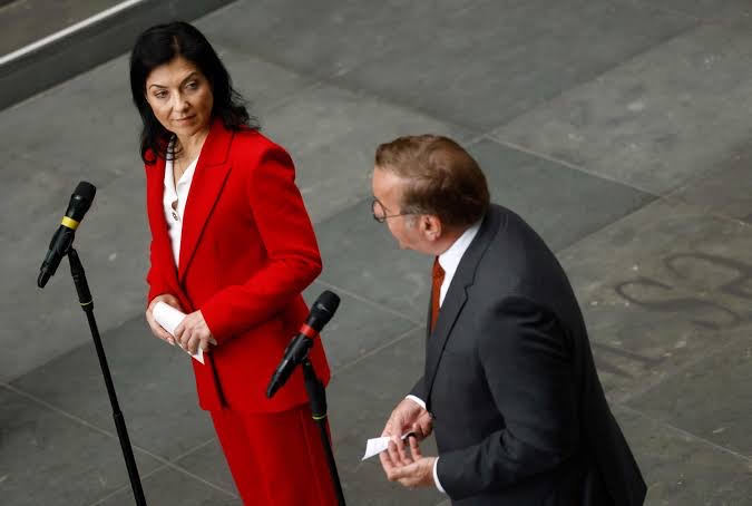 Katherina Reiche in a red suit at microphones, speaking with a colleague in a hall